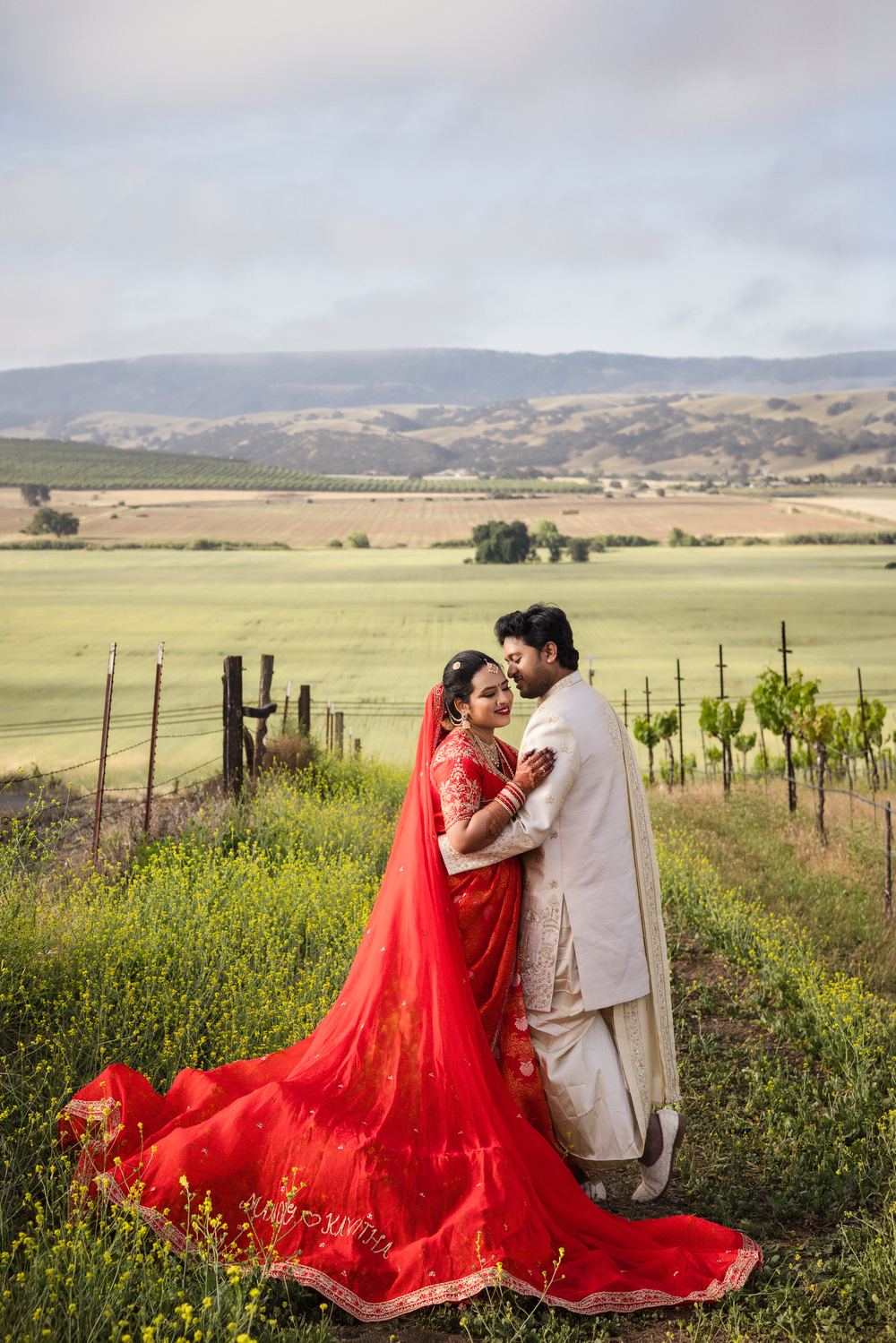 A bride in red with a long embroidered train, held close in the vineyard hills