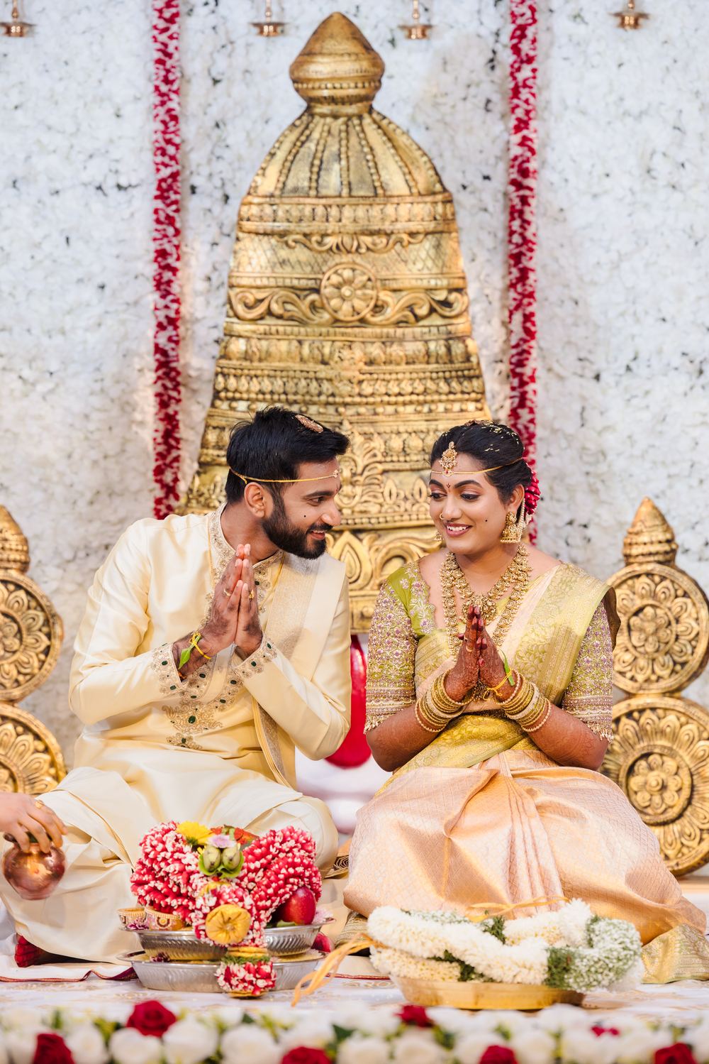 A couple in gold and green before a gilded mandap, hands folded in prayer