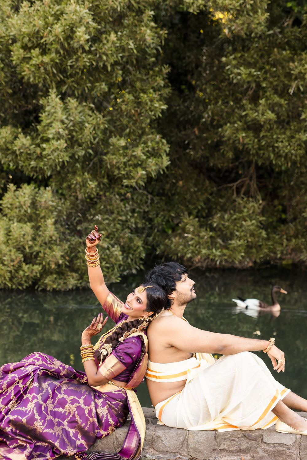 A bride in purple silk mid dance pose beside a pond, a swan on the water