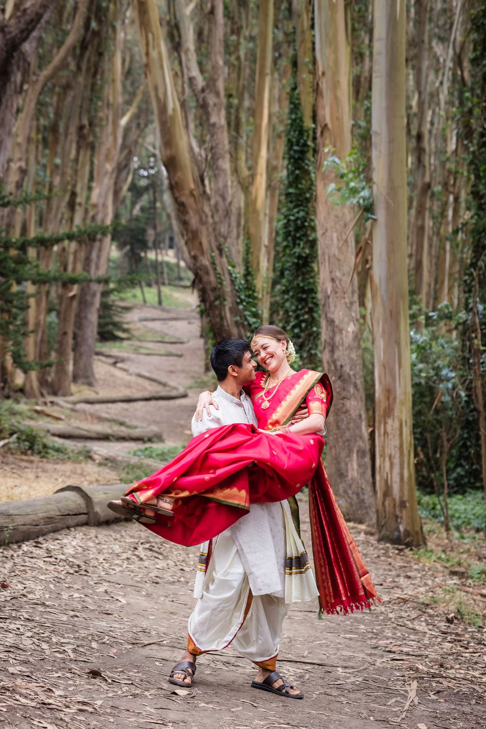 A groom carrying his bride down a eucalyptus path, both laughing